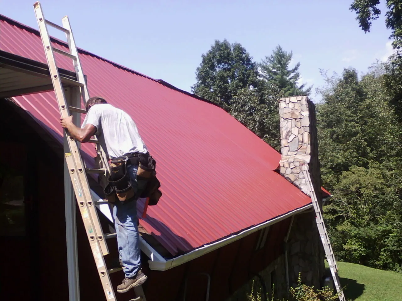 Roofer on a ladder beside a red metal roof and stone chimney — Soffit & Fascia Repair by Roofing Brothers in Vincent